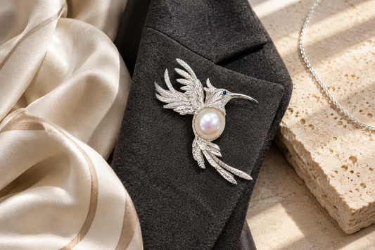 HEWREN handcrafted sterling silver hummingbird brooch styled on a tailored charcoal blazer lapel beside pure silk drape and silver chain on limestone
