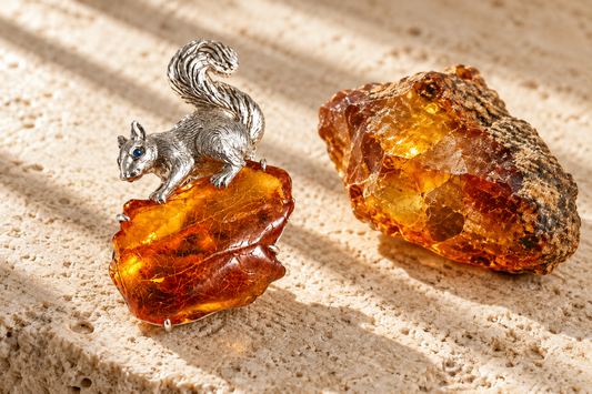 HEWREN handcrafted sterling silver squirrel brooch clutching golden Baltic amber beside a raw rough amber specimen on travertine stone